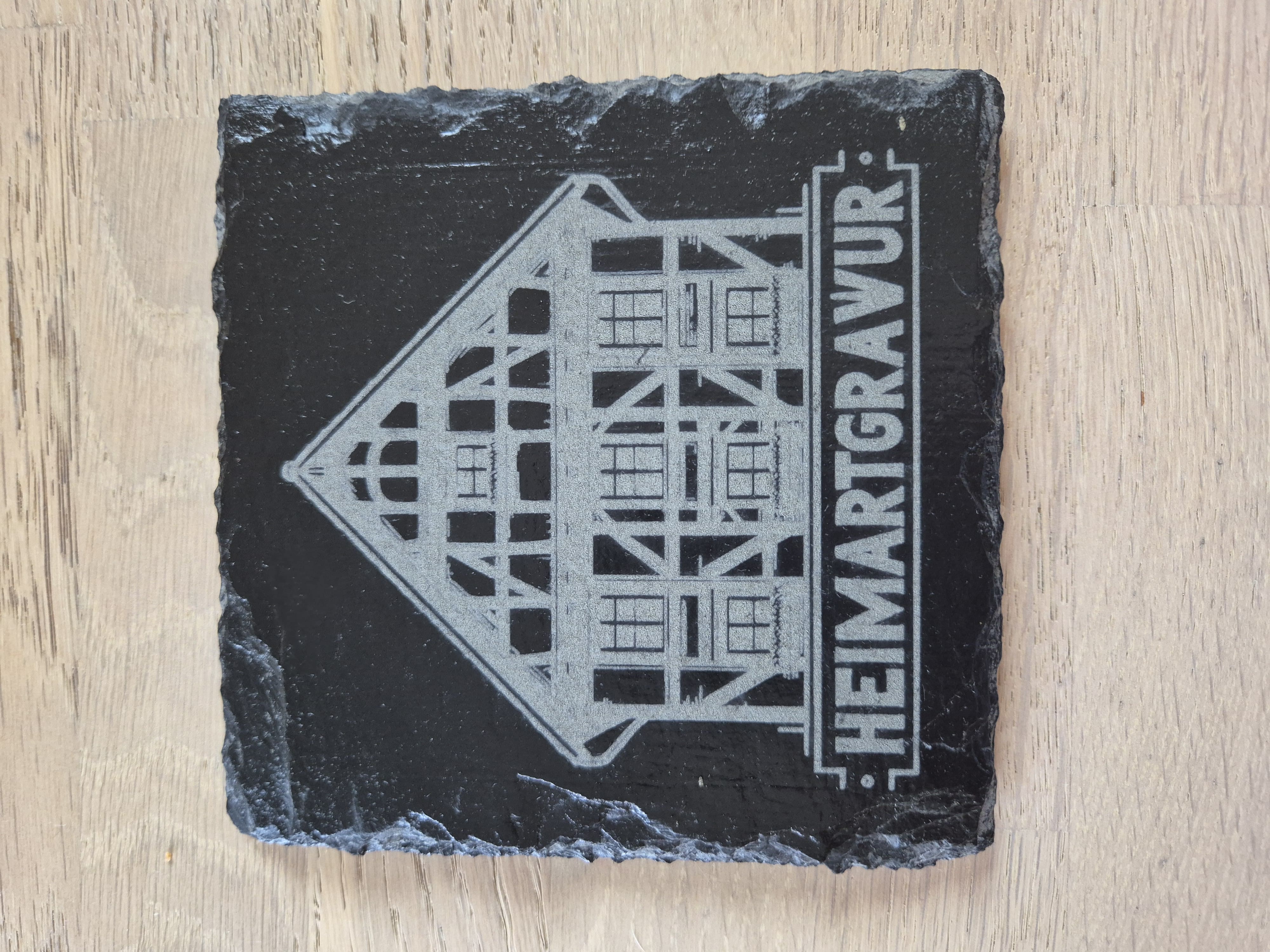Graviertes Logo von HeimArtGravur auf einer 13x13 cm Schieferplatte. 